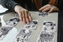 A person reaching for a card from a set of black and white illustrated game cards laid out on a table. The cards feature intricate designs and varying themes, encouraging engagement and play.