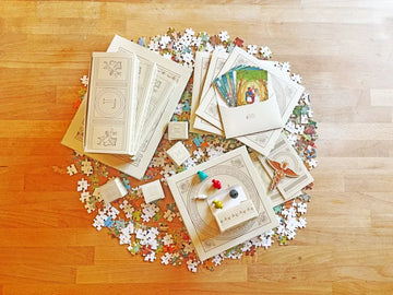 Components of Pandora's Legacy, including jigsaw pieces, meeples, instruction cards, and boxes with stylistic art arranged neatly in a circle on a wooden floor