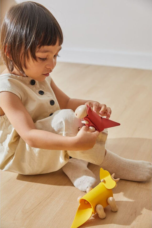 A young child wearing a beige outfit sits on a wooden floor, focused on assembling the DIY Tyrannosaurus kit by Plan Toys. The child is holding a red component of the dinosaur while a yellow piece lies nearby, showcasing the creative play and hands-on learning experience the kit offers.