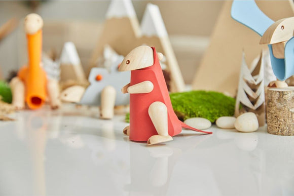 A DIY kit featuring a colorful wooden Tyrannosaurus figure by Plan Toys, displayed among other playful wooden animals and natural elements like moss and stones, perfect for creative play and learning.