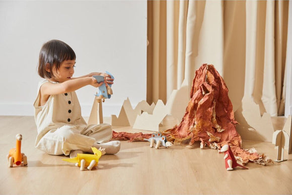 A young child sits on the floor playing with the DIY Tyrannosaurus kit by Plan Toys. They are holding a blue dinosaur as part of a creative play setup that includes various colorful dinosaur figures and a paper mountain in the background, promoting imaginative play.