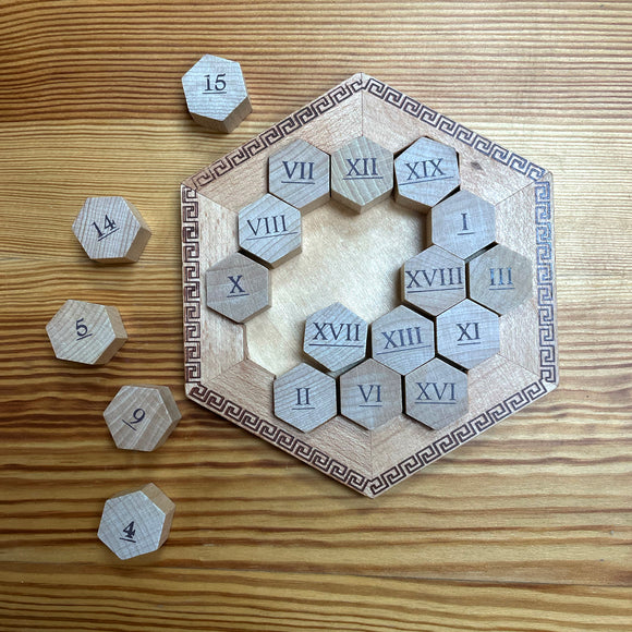 The Hypatian Enigma by True Genius is a mechanical puzzle featuring a hexagonal wooden base with intricate Greek patterns. The puzzle includes multiple hexagonal pieces, each engraved with Roman numerals ranging from I to XV. The pieces are arranged in a way that invites players to explore various combinations and solutions, providing a challenging and engaging experience for puzzle enthusiasts.