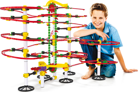 A young boy sits next to a colorful Skyrail Ottovolante elevator by Quercetti. The DIY kit features multiple winding tracks in red, yellow, and green, creating an intricate elevator system. The boy is smiling, highlighting the fun and engaging nature of the product, suitable for imaginative play and learning.