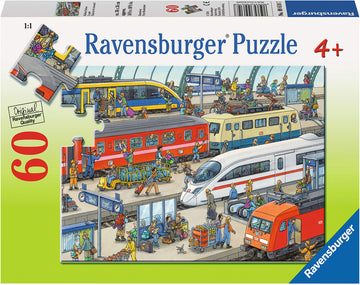 A colorful 60-piece jigsaw puzzle by Ravensburger featuring a lively railway station scene. The image shows various trains, busy travelers, and railway staff, capturing the hustle and bustle of a train station with detailed illustrations and vibrant colors, suitable for ages 4 and up.
