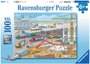 A colorful jigsaw puzzle titled 'Construction at the Airport' by Ravensburger. The image features a busy airport scene with various construction vehicles, workers in safety gear, airplanes on the runway, and an airport terminal in the background. It highlights the hustle and bustle of construction activity, showcasing trucks, cranes, and machinery working together. Designed for ages 6 and up.