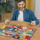 A person smiling while assembling the Peanuts Moments jigsaw puzzle on a wooden table. The puzzle features colorful illustrations of Peanuts characters, scattered pieces surround the partially completed puzzle. The setting includes indoor plants and warm lighting, creating a cozy atmosphere.