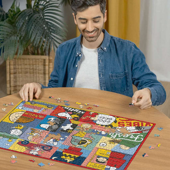 A person smiling while assembling the Peanuts Moments jigsaw puzzle on a wooden table. The puzzle features colorful illustrations of Peanuts characters, scattered pieces surround the partially completed puzzle. The setting includes indoor plants and warm lighting, creating a cozy atmosphere.