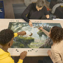 A vibrant jigsaw puzzle titled 'The Forest Dragon Awakes' manufactured by Ravensburger is being assembled by three people at a wooden table. The puzzle depicts a fantasy scene with a dragon among lush greenery, a serene forest, and deer. Various puzzle pieces are scattered around them.