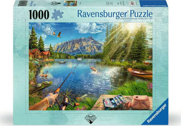 Ravensburger jigsaw puzzle titled 'Life at the Lake' featuring a serene lakeside scene. The image depicts a tranquil lake surrounded by lush trees, a few fishing boats, and cabins. A person is seen fishing, and ducks are swimming in the water under a bright blue sky with fluffy clouds.