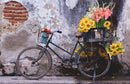A rustic bicycle parked against a textured wall, adorned with a wooden crate overflowing with bright yellow sunflowers, pink and white gladiolus flowers, adding a vibrant touch to the scene.