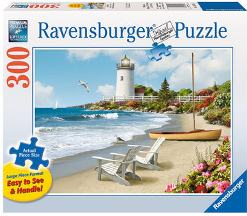 The Sunlit Shores 300 pc Large Format Puzzle by Ravensburger features a serene beach scene. It captures a picturesque view with a lighthouse, sailboat, and beach chairs by the shore, surrounded by colorful flowers and a calm ocean. The puzzle pieces are designed in a large format for easy handling and visibility, making it accessible for all ages.