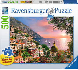 Ravensburger Positano Jigsaw Puzzle featuring a stunning view of the coastal town at sunset. This 500-piece puzzle showcases colorful buildings, vibrant flowers, and sailboats on the calm sea. The large format pieces are designed for easy handling and enhanced visibility.