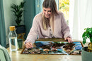 A young woman sitting at a table, working on a 1000-piece Harry Potter jigsaw puzzle by Ravensburger. The puzzle features beloved characters from the series and is nearly completed. A glass of water and a potted plant are visible on the table, creating a cozy atmosphere.