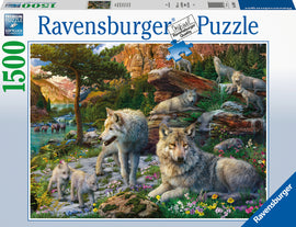 Ravensburger Wolf Wilderness 1500-piece jigsaw puzzle featuring a beautiful scene of wolves in a lush wilderness setting with mountains, trees, and a flowing stream. The high-quality image showcases playful wolf cubs, a watchful adult wolf, and other wildlife elements in a vibrant natural environment.