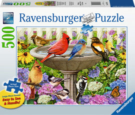 A colorful jigsaw puzzle box by Ravensburger featuring a vibrant scene of various birds, including a cardinal, bluebird, and others, gathered around a birdbath surrounded by flowers. The box highlights a '500' piece count and emphasizes the large piece format designed for easy handling.