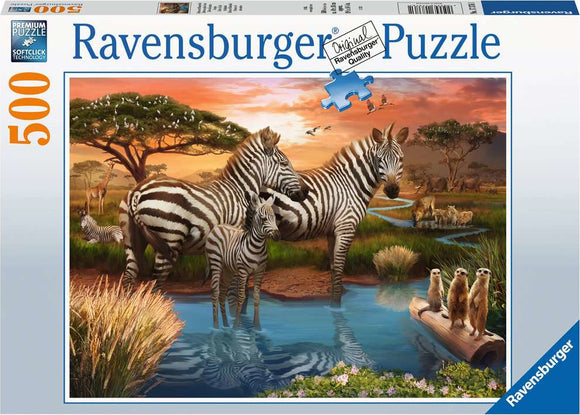 A 500-piece jigsaw puzzle titled 'Zebras at the Waterhole' by Ravensburger. The image features zebras drinking water at a serene waterhole surrounded by lush greenery, with meerkats on a log and giraffes in the background under a vibrant sky.
