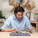 A young man with dark hair is smiling while holding puzzle pieces above a colorful jigsaw puzzle on a table. The puzzle features a vibrant nature garden scene with various plants and flowers. In the background, decorative shelves hold books and a vase with tulips.
