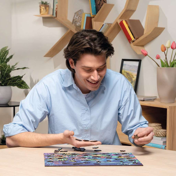 A young man with dark hair is smiling while holding puzzle pieces above a colorful jigsaw puzzle on a table. The puzzle features a vibrant nature garden scene with various plants and flowers. In the background, decorative shelves hold books and a vase with tulips.