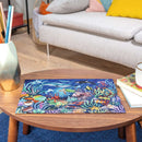 A beautifully illustrated 500-piece jigsaw puzzle titled 'WOOD: Under the Sea' by Ravensburger, featuring vibrant underwater scenery with colorful fish, sea turtles, and coral reefs, displayed on a wooden coffee table. Surrounding elements include a geometric vase with colored pencils, a cozy sofa, and books stacked nearby, creating a warm and inviting atmosphere.
