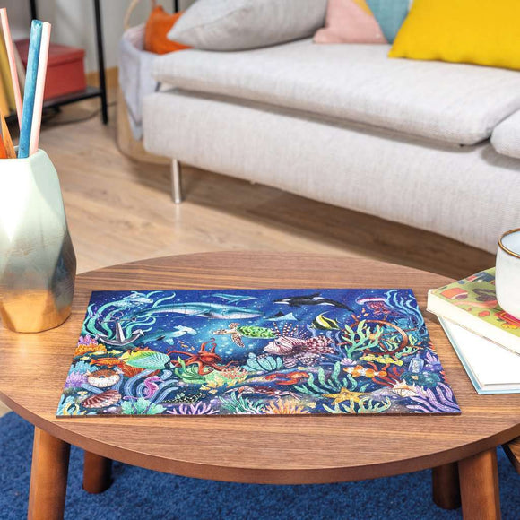 A beautifully illustrated 500-piece jigsaw puzzle titled 'WOOD: Under the Sea' by Ravensburger, featuring vibrant underwater scenery with colorful fish, sea turtles, and coral reefs, displayed on a wooden coffee table. Surrounding elements include a geometric vase with colored pencils, a cozy sofa, and books stacked nearby, creating a warm and inviting atmosphere.