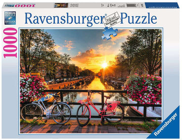 Ravensburger 1000-piece jigsaw puzzle featuring a picturesque scene in Amsterdam. The image showcases a canal with bicycles parked by a wooden railing, surrounded by colorful flowers and trees, under a stunning sunset sky.