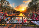 A picturesque jigsaw puzzle featuring two bicycles, one white and one pink, parked by a canal in Amsterdam at sunset. The scene includes colorful flowers in the foreground and the tranquil water reflecting the sunset hues.