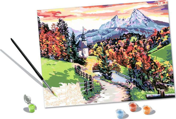 CreArt Beautiful Bavaria DIY kit showcasing a picturesque landscape of vibrant autumn colors, with a winding path, church steeple, and majestic mountains in the background. The kit includes a paintbrush, paint pots, and a canvas featuring outlines for easy painting.