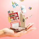 A delicate hand holds a beautifully crafted DIY Childhood Toy House by Robotime. The toy features a vibrant pink display shelf filled with miniature items like a teddy bear, books, and colorful decorations. A playful polar bear figurine and colorful blocks float above the house, with a charming 'Lovely Room' label at the base, showcasing creativity and intricate design.