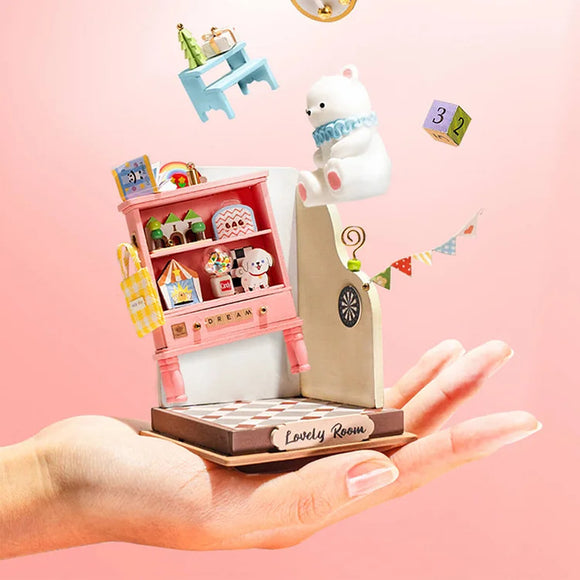 A delicate hand holds a beautifully crafted DIY Childhood Toy House by Robotime. The toy features a vibrant pink display shelf filled with miniature items like a teddy bear, books, and colorful decorations. A playful polar bear figurine and colorful blocks float above the house, with a charming 'Lovely Room' label at the base, showcasing creativity and intricate design.