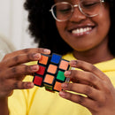 A close-up of a young person with curly hair and glasses joyfully solving a Rubik's Cube - 3x3 from Spin Master, showcasing their engaging focus and colorful puzzle.