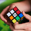 A close-up of a person's hands holding a Rubik's Cube 3x3 puzzle from Spin Master. The cube features vibrant colors: red, white, blue, green, orange, and yellow on each side, showcasing the classic design of a mechanical puzzle. The background is blurred, focusing on the intricate details of the Rubik's Cube.
