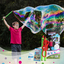 A joyful child wearing glasses holds a pair of green bubble wands, creating a gigantic rainbow-hued bubble in a park. Next to him, the WOWmazing Grab-n-Go Giant Bubble Kit package and its components are visible, showcasing a fun outdoor activity for kids.