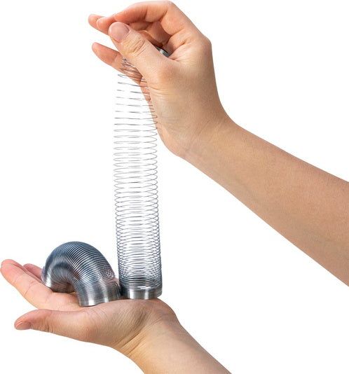 A Mini Sproing toy by Schylling being held in two hands. The toy is a shiny metal spring, partially coiled and partially extended, showing its flexible design. One hand supports a small curled section of the spring while the other hand lifts the elongated section, demonstrating its ability to stretch and compress for fun and engaging play.