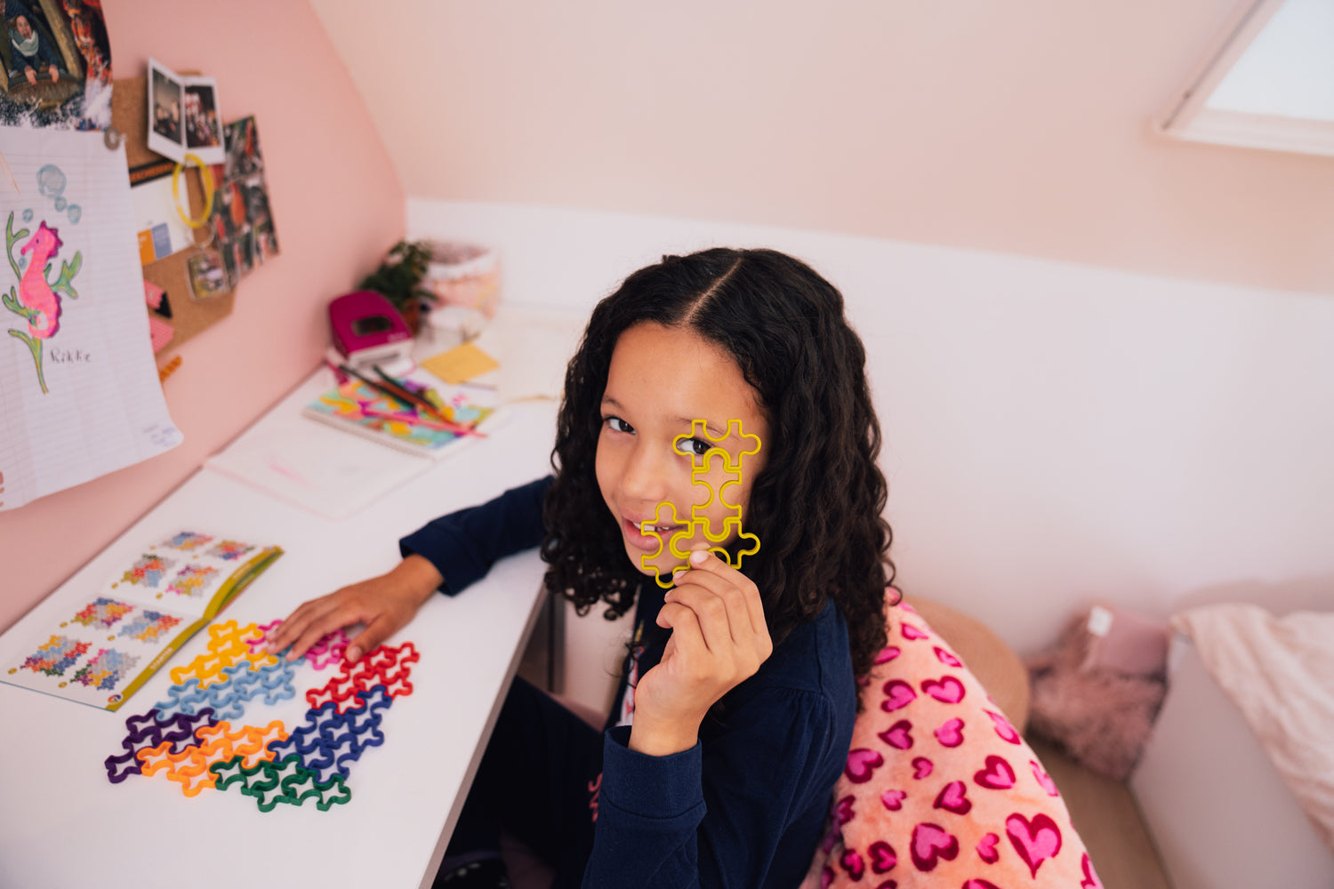 A young girl smiles while holding up two colorful jigsaw puzzle pieces in front of her face. She is seated at a desk covered with notebooks, colorful papers, and additional puzzle pieces. The room features pastel walls and a cozy atmosphere.