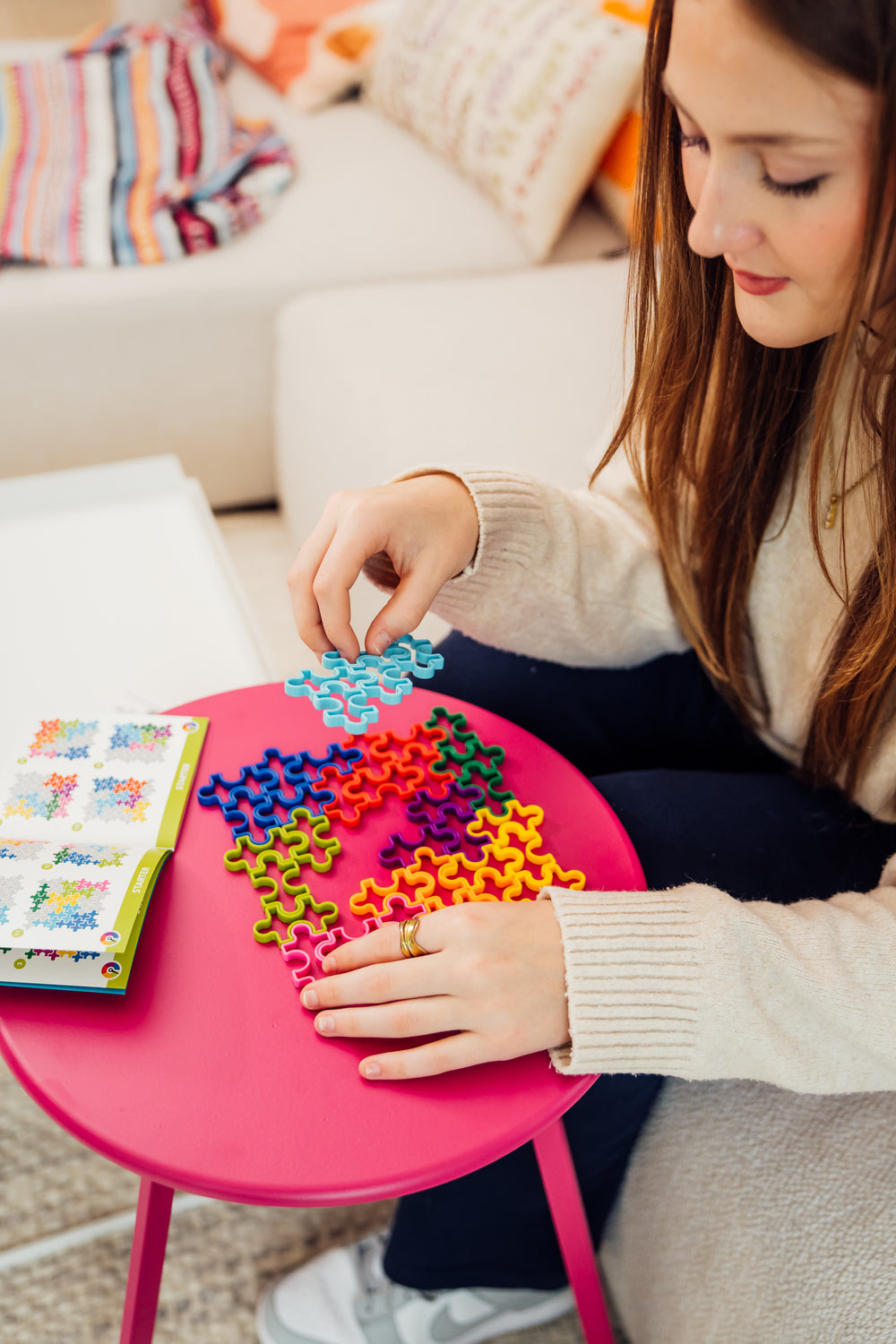 A person assembling the IQ Jigsaw puzzle by Smart Toys & Games. The puzzle pieces are colorful, displayed on a bright pink table, with instruction sheets visible, showcasing the creative and mechanical nature of the puzzle.