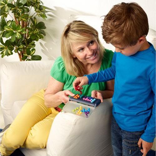 A mother and her young son sit together on a couch, engaged in playtime with the IQ Fit mechanical puzzle from SmartGames. The mother is happy as she helps her son place colorful puzzle pieces in a compact tray, promoting cognitive skills and teamwork. A green plant is visible in the background, creating a cozy atmosphere.
