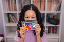 A child with curly hair smiling while holding the IQ Waves mechanical puzzle toy, which features colorful, interlocking pieces in a compact storage case. The background shows a cozy room with shelves filled with books.