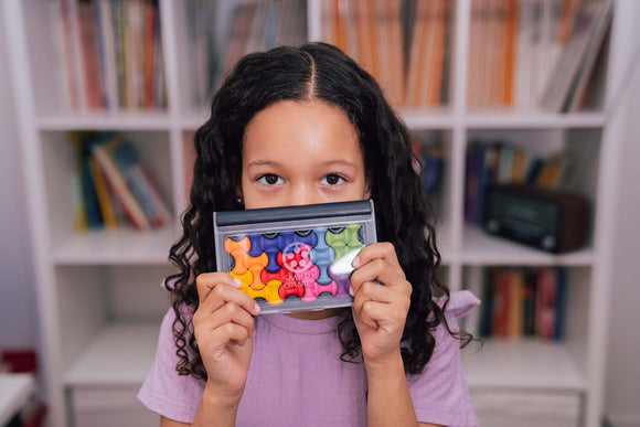 A child with curly hair smiling while holding the IQ Waves mechanical puzzle toy, which features colorful, interlocking pieces in a compact storage case. The background shows a cozy room with shelves filled with books.