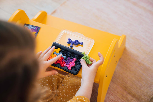 A child engaged in the IQ Waves mechanical puzzle by Smart Toys & Games, manipulating colorful, uniquely shaped pieces. The puzzle pieces are housed in a black tray with a clear lid on a bright yellow table.