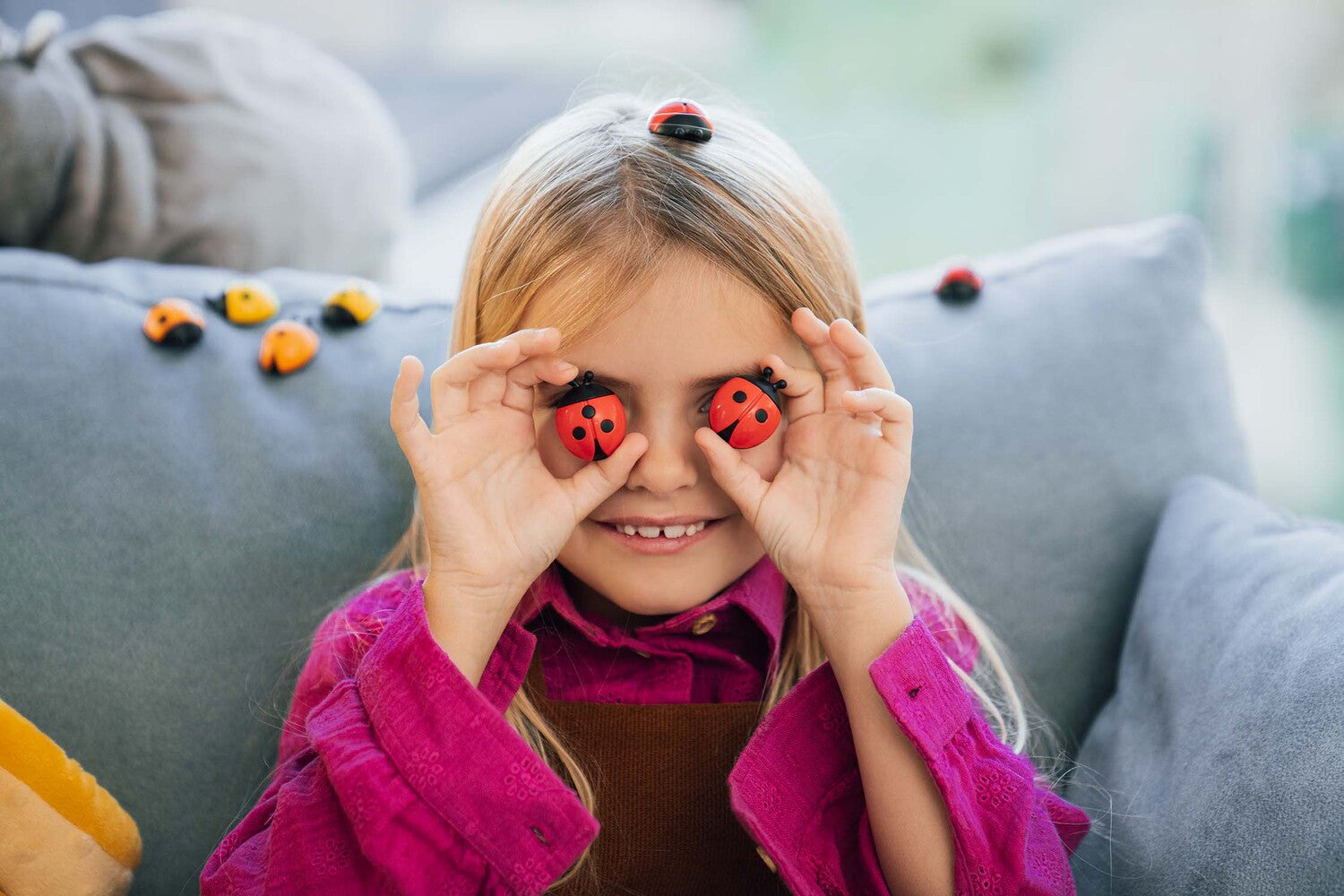 A young girl with long blonde hair smiles while holding colorful ladybug-shaped toys over her eyes. She wears a bright pink shirt and has playful ladybug figures on her head and around her on the couch. The setting is cozy, with a soft background.