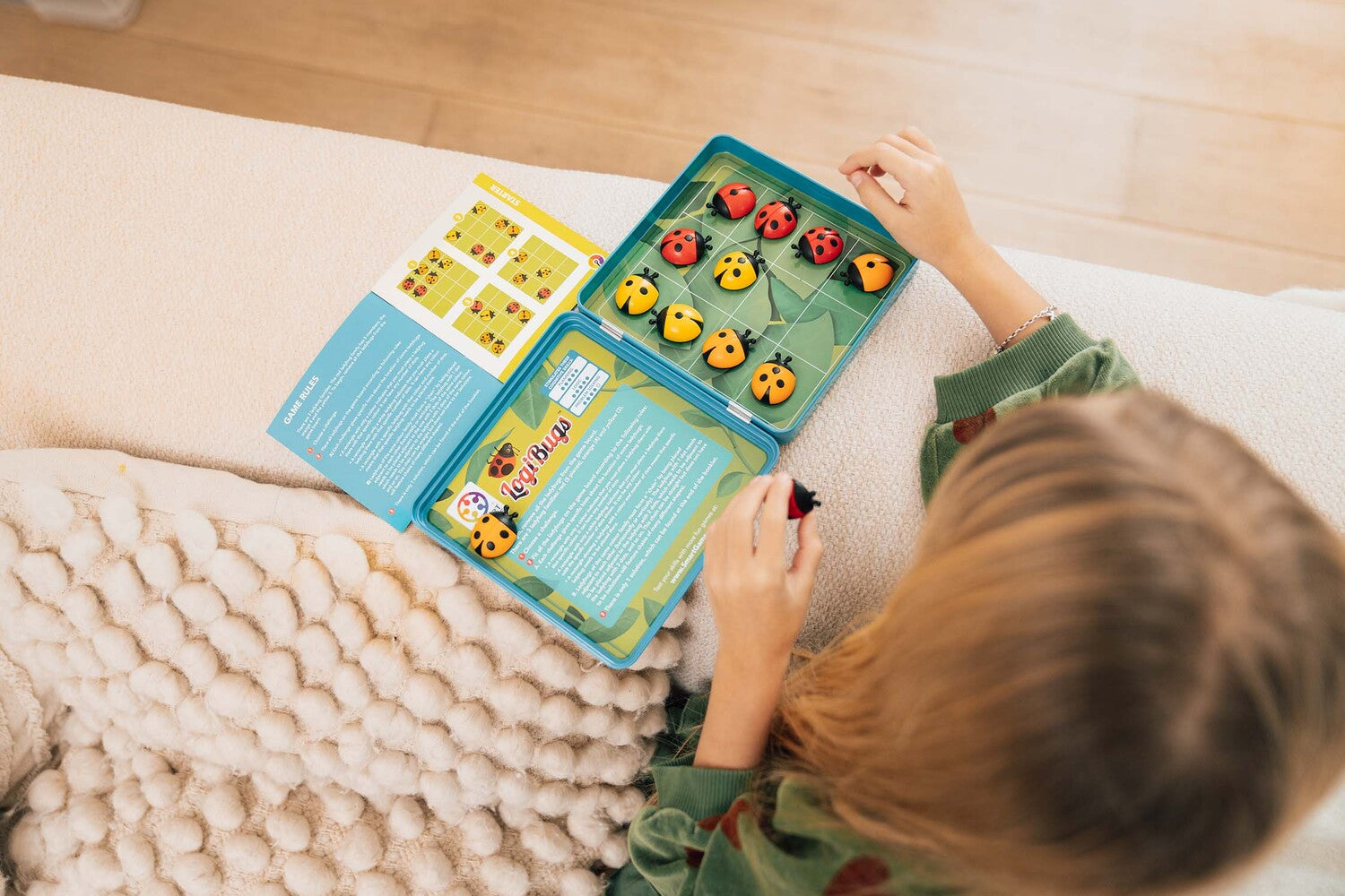 A child plays with LogiBugs, a mechanical puzzle set by Smart Toys & Games. The puzzle features colorful ladybug pieces arranged on a green board. An open instruction booklet is visible, providing game rules and challenges. The child appears engaged in solving the puzzle, with a cozy blanket in the background.