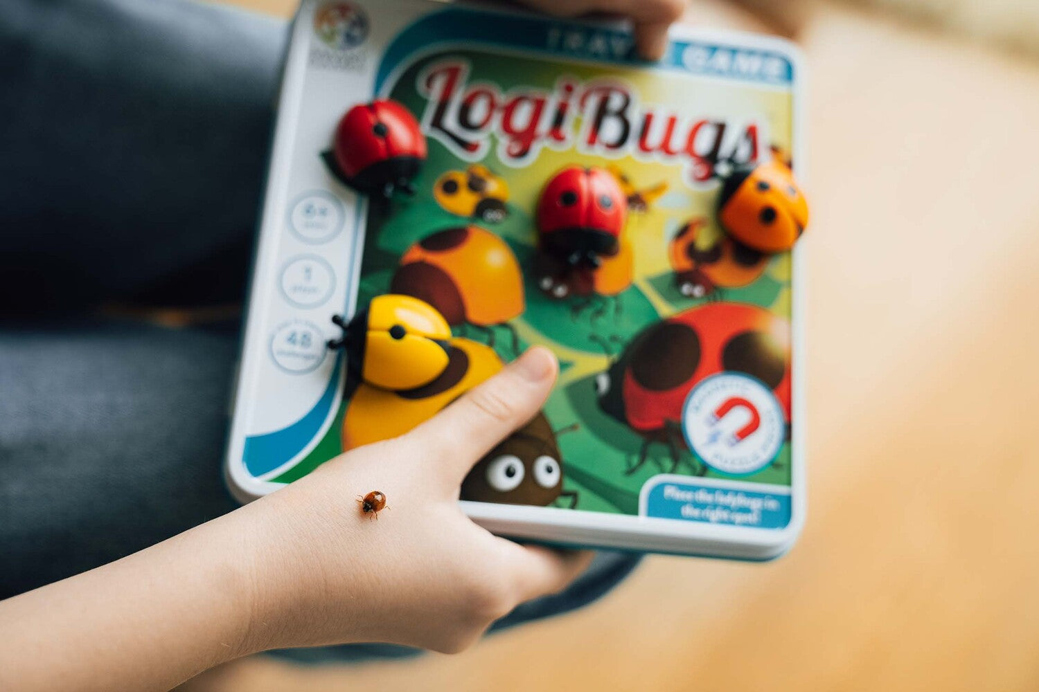 A child holds a game called LogiBugs by Smart Toys & Games. The game features colorful mechanical bugs that must be placed correctly on a vibrant game board with various insect designs. A ladybug is resting on the child's hand, adding a playful element to the scene. The game is designed for ages 4 and up and is suitable for one or more players, containing 48 challenges.