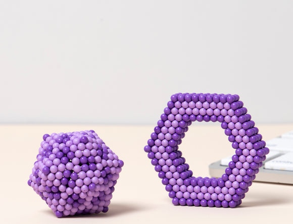 The Speks Elements: Cumulus features two colorful magnetic construction pieces in shades of purple. One is a compact hexagonal shape, while the other resembles an abstract form, both made of small magnetic beads. They are presented against a neutral background and a white keyboard is partially visible in the image.
