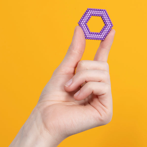 A hand holding a purple hexagonal piece made of small magnetic balls, set against a bright orange background. The hexagon showcases a unique geometric design, inviting tactile interaction.