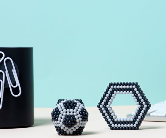Speks Elements: Lunar features a colorful collection of magnetic balls, shaped into two distinct structures: a spherical form and a hexagonal shape. The objects are set against a soft blue background, with a sleek black container holding paper clips in the corner, creating a modern and minimalist workspace aesthetic.
