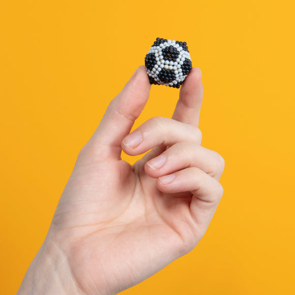 A hand holding a small, black and white spherical fidget toy resembling a soccer ball, set against a bright yellow background. The toy's intricate design features small beads that create a textured surface, making it visually appealing and satisfying to touch.