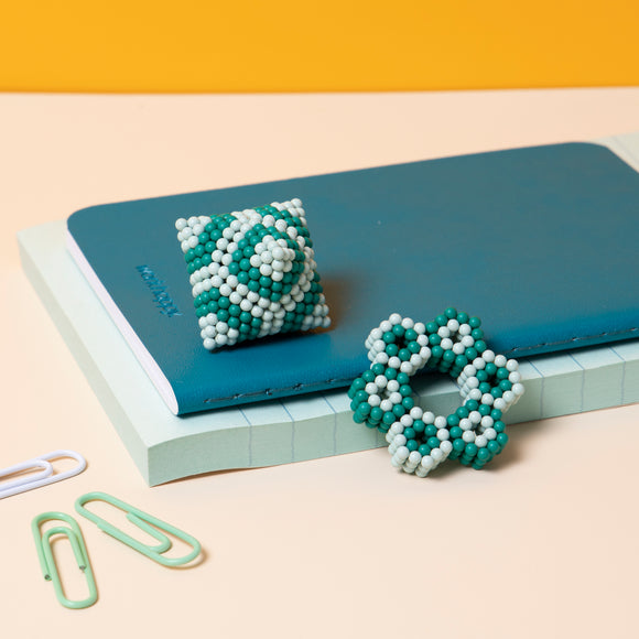 A colorful set of Speks Elements: Marine, featuring a decorative fidget toy in intricate designs made of teal and white magnetic beads. The items are displayed on a layered background of notebooks and paper clips, emphasizing their artistic and playful nature.