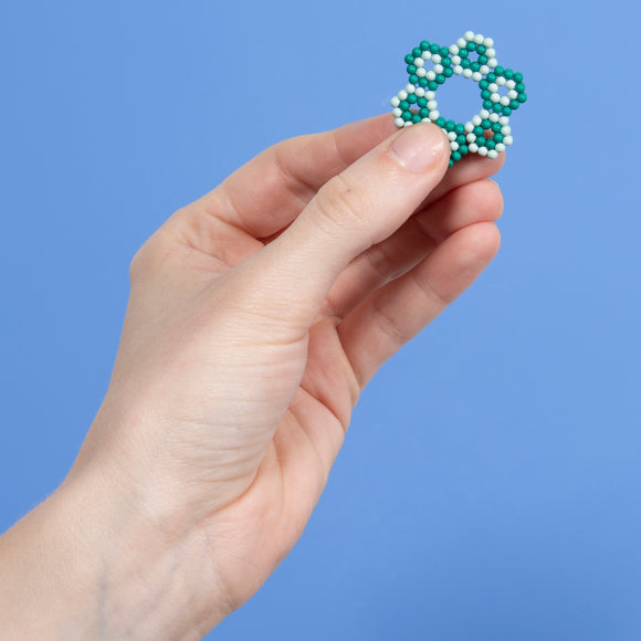 A person's hand holds a small, intricate Green and White Speks Elements: Marine piece, featuring a unique floral design. The background is a solid light blue, emphasizing the vibrant colors of the toy.