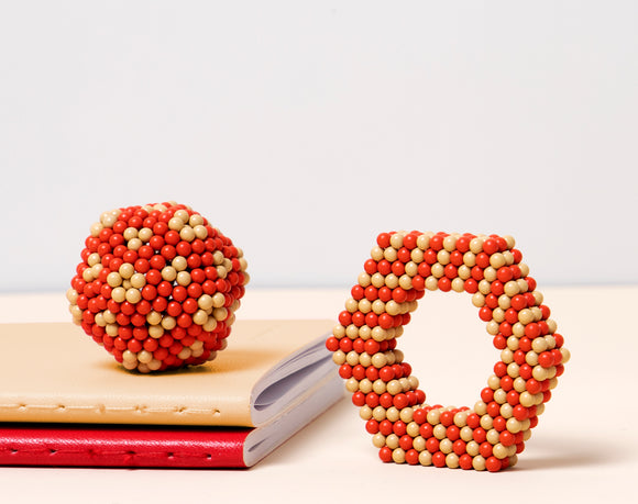 Speks Elements: Solar, a creative and colorful magnetic building set featuring a spherical shape and a hexagonal structure made of small, spherical magnets in red and beige. The pieces are showcased on a light-colored surface, along with partially visible notebooks in various colors, promoting a playful and inspiring atmosphere for creativity and tactile engagement.