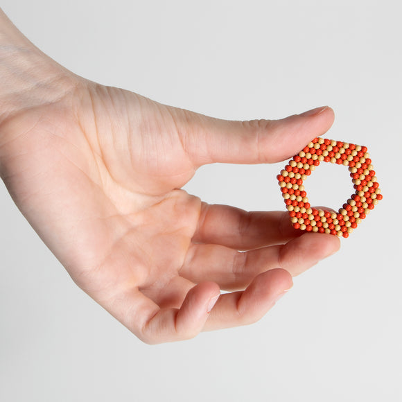 A person holds a hexagonal shape made of small magnetic balls in shades of orange and beige. The design is intricate, showcasing the versatile construction possibilities of the Speks Elements: Solar product.